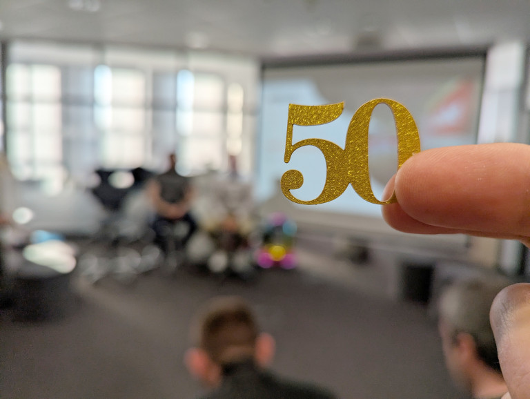 Die Zahl 50, aus goldenen Papier, werden von zwei Fingern gehalten. Im Hintergrund, verschwommen, drei personen die vor eine Leinwand sitzen um eine Präsentation zu halten.