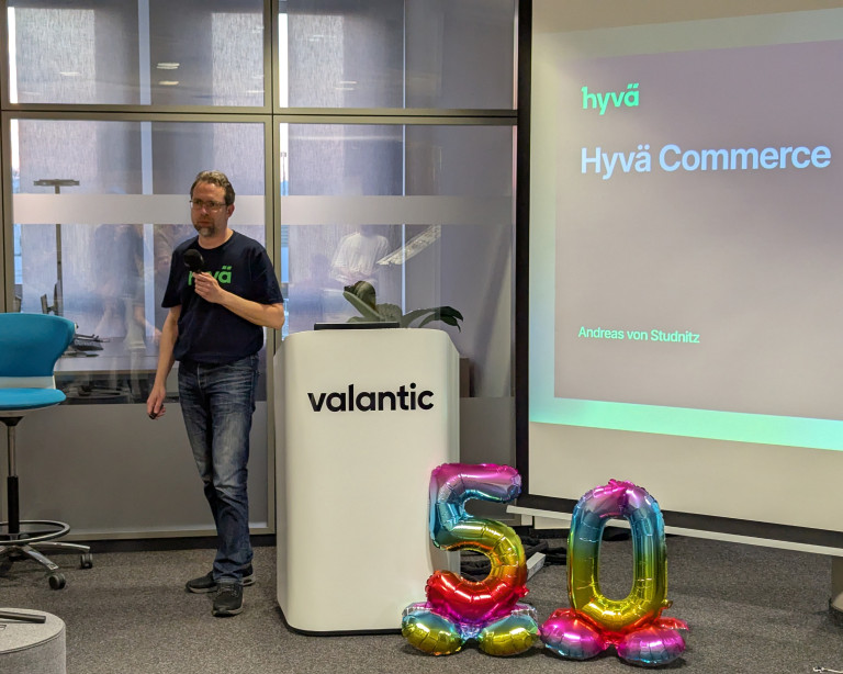 Andreas von Studnitz (Senior Operations and Magento Architect, Hyvä) stegt links von einer Leinwand auf dem seine Präsentation zu Hyvä Commerce dargestellt wird.